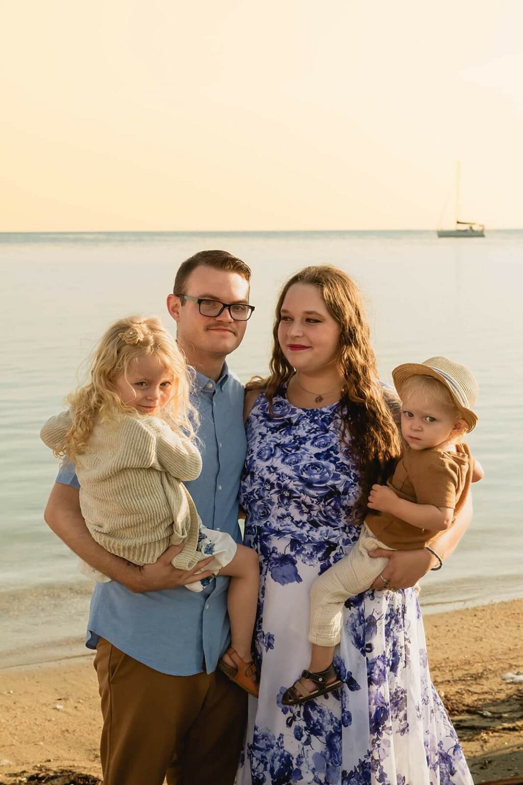 A family of four on a serene beach. The mother is wearing a long white dress with cobalt blue flowers and has long hair. She is holding their sweet son and is embracing her husband who has a blue shirt and khaki pants and is holding their daughter.
